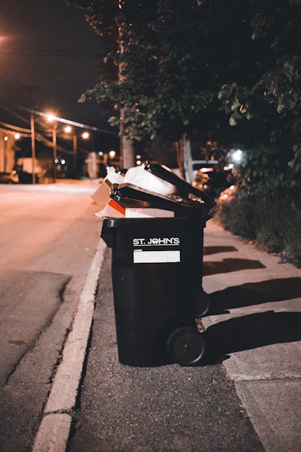 A black wheeled rubbish bin positioned on the pavement near the edge of a street at night. The bin has a white label with the name 'St. John’s' and contains various discarded items including paper, cardboard, and other waste materials. Streetlights cast a warm glow along the deserted road, illuminating the scene and creating shadows from the bin and nearby foliage. In the background, there are trees, utility poles, and faintly visible parked cars, indicating a quiet residential or suburban area. The surrounding environment appears clean and organized, with the bin placed conveniently for drop-off of waste in line with private waste management services like those offered by Rubbish Collection Hampstead, which may provide alternative rubbish collection solutions beyond local authority disposal options. Overall, the scene emphasizes the typical setting for refuse collection and rubbish removal activities in an urban or suburban environment during nighttime hours, highlighting the importance of proper waste disposal at a local level.