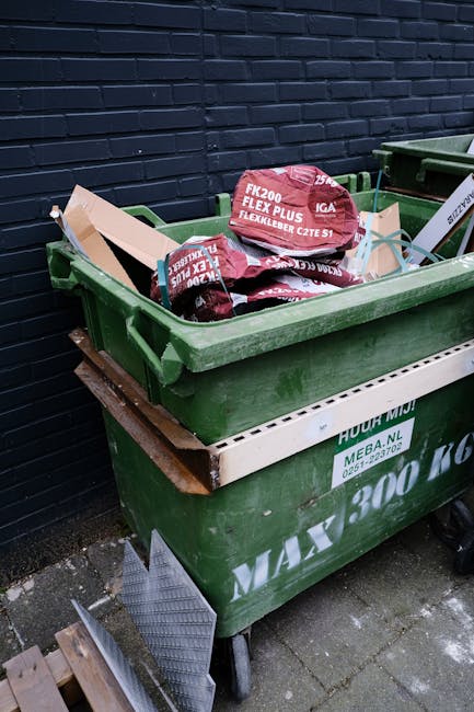 A large green wheeled skip bin positioned outdoors on a paved surface against a dark brick wall, filled with various discarded bags of garden materials and landscaping supplies. The bags are predominantly red with some white text, indicating they contain flexible rubber or similar materials. The skip shows signs of weathering and is stacked on a wooden pallet, suggesting it is used for private waste disposal or alternative rubbish removal services. In the background, there is a second, similar green skip to the right, partially visible. The scene is illuminated by natural light, emphasizing the textures of the bags, bin, and brick wall, aligning with the context of professional rubbish collection and waste management related to business or shop premises in Hampstead.