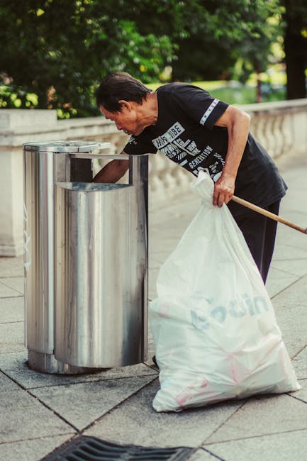 A middle-aged woman with short, dark hair, dressed in a black t-shirt and dark pants, is seen bending over to dispose of a large white garbage bag into a stainless steel, cylindrical litter bin with a vertical slot on the top, situated on a paved sidewalk. The woman holds a wooden broom in her right hand, resting against her hip, and appears to be engaged in rubbish collection or waste disposal. The scene is set outdoors, with lush green foliage and trees visible in the background, along with a stone or concrete balustrade that runs parallel to the sidewalk. The environment suggests a public park or residential area, and the overall atmosphere is calm and orderly, consistent with a private or community rubbish removal service like those offered by Rubbish Collection Hampstead. The stainless steel bin has a smooth, reflective surface, contrasting with the textured, light gray paving stones beneath. Natural daylight illuminates the scene, emphasizing the clean, organized presentation of waste management activities.