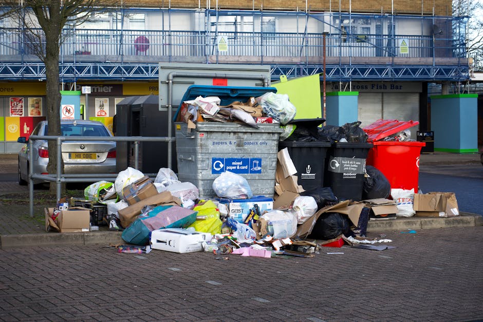 A large pile of mixed rubbish and waste bags, including cardboard boxes, plastic bags, paper, and packaging materials, is overflowing from black and gray recycling and waste bins placed on a paved sidewalk. The waste is scattered around the bins, with some items spilling onto the surrounding brick paving area. Behind the bins, there is a blue construction scaffold covering a building, with green and white safety signs visible on the upper level. To the left, a small silver car is parked close to a metal railing that runs along the edge of the sidewalk. The overall scene depicts an uncollected waste accumulation, illustrating the need for efficient rubbish removal services, such as those provided by Rubbish Collection Hampstead, for managing excess waste outside standard collection points, especially in busy urban areas like Hampstead.