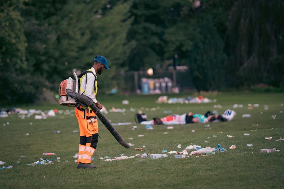 A sanitation worker dressed in high-visibility orange and black trousers, a white jacket with reflective strips, and a blue cap, is using a leaf blower to clear litter from a grassy park area during daylight. The worker is positioned on the left side of the image, with a large, orange backpack-style leaf blower strapped to their back, and their gaze directed downward at the ground. In the background, there are two figures lying on the grass, surrounded by various types of trash, including plastic bottles, paper, and debris scattered across the lawn. The scene is set in a park with dense green trees and bushes in the backdrop, and a small structure or waste station is visible behind the figures. The lighting appears natural, possibly during late afternoon or early evening, highlighting the effort of waste management in outdoor public spaces to maintain cleanliness and environmental awareness. This scene emphasizes the importance of professional rubbish removal and cleanup services, such as those provided by Rubbish Collection Hampstead, especially after public events or gatherings, to support effective alternative waste handling and on-site clearance.