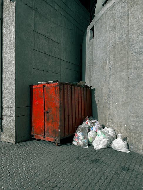The image depicts a narrow urban alleyway cluttered with various discarded waste materials, primarily large black plastic bags and a bulky, weathered grey garbage sack positioned in the foreground, standing upright on a worn, dark asphalt surface. Behind these, a pile of cardboard boxes, some flattened and others partially intact, along with white plastic bags and miscellaneous rubbish, are stacked against a makeshift barrier at the end of the alley. The surrounding environment includes the facades of adjacent buildings on each side: the left with a rusty metal door and graffiti-covered wall, and the right with a dark grey wall featuring vertical metal bars and a window with security bars. Bare tree branches extend overhead, casting a sparse, diffuse light across the scene, which emphasizes the grim, neglected atmosphere typical of private waste handling efforts in urban settings. Occasionally, small debris like a plastic bottle and bits of paper are visible scattered on the ground, highlighting the cluttered and unmaintained state of this alley, consistent with independent rubbish collection sites managed by local waste services such as Rubbish Collection Hampstead.