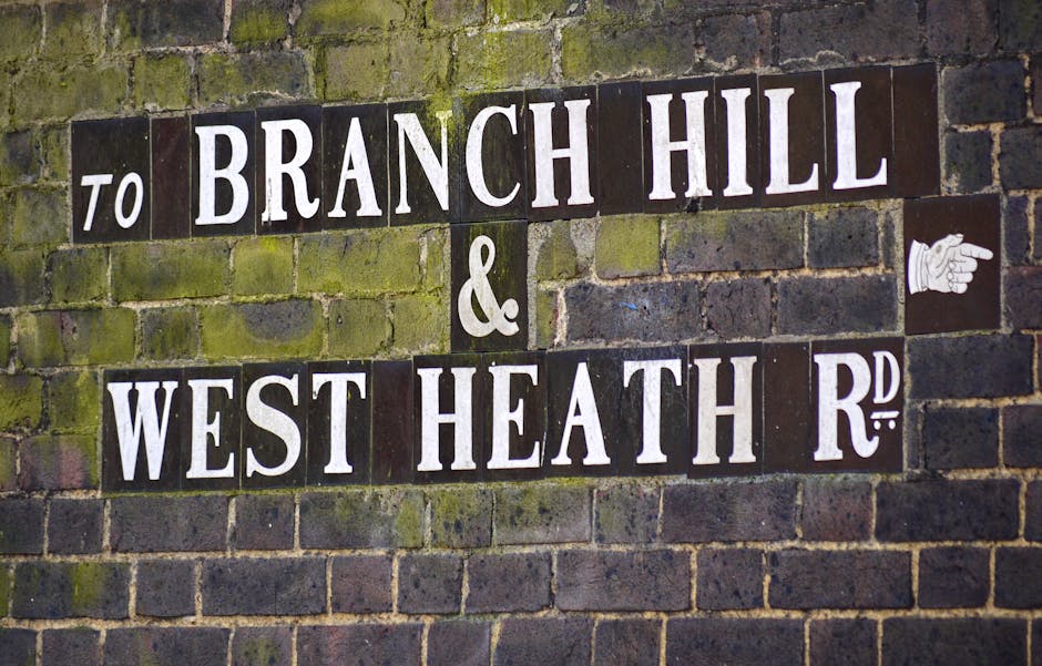 A close-up of a traditional street sign mounted on an aged brick wall, indicating directions to Branch Hill and West Heath Road. The sign features black ceramic tiles with white uppercase lettering, with some tiles showing slight weathering and moss growth, suggesting outdoor placement. To the right of the sign, there is a small white painted hand pointing towards the direction, possibly part of historic street signage. The brick wall behind the sign has a mottled appearance with dark gray, brown, and greenish moss patches, indicating exposure to outdoor elements. The environment appears to be in a residential or historical urban area, with the sign positioned slightly to the right in the image, suggesting it is mounted on the exterior of a building or wall adjacent to a pathway or street. The overall scene captures a quiet, possibly older part of town where traditional street signage remains in place, reflecting the local area's character and history, which is relevant to the context of local waste management and street-related signage for residents and visitors.