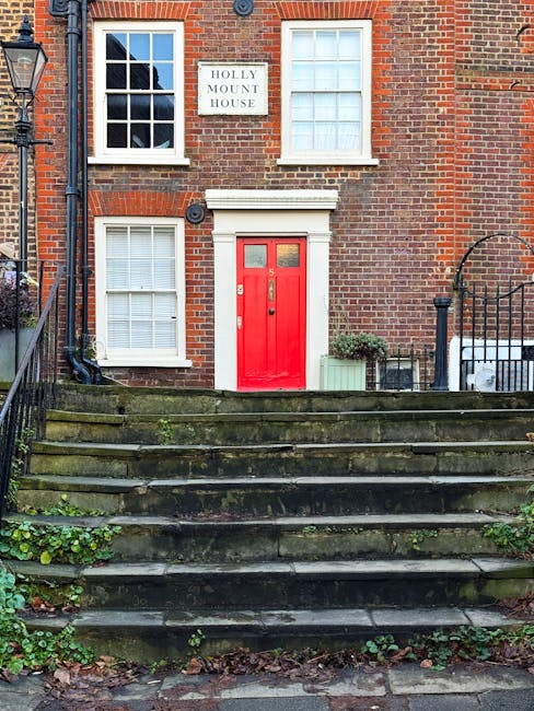 The image depicts a traditional brick residential building with a set of weathered stone steps leading up to a bright red front door. The door features two small rectangular glass panels at the top and is framed by a white trim. To the left of the door are two white-framed windows with multiple panes, one positioned on the ground floor and the other directly above on the first floor, both fitted with closed white blinds. On the right side of the building, a decorative black wrought-iron fence borders a small front garden area, which contains a green rectangular planter with foliage and a small bush. A black gutter pipe runs vertically down the left side of the building, near a vintage-style street lamp attached to the left edge of the image. A white sign with black lettering reading 'HOLLY MOUNT HOUSE' is affixed between the two upper windows, indicating the property is part of a private residence or collection of homes. The stone steps are mossy with patches of greenery growing between the crevices, contributing to an aged appearance, and fallen leaves are scattered at the bottom of the stairs, suggesting an outdoor setting typical of an English neighbourhood. This scene subtly relates to the context of on-site waste management or private rubbish collection services, as it shows a residential facade potentially serviced by independent waste collection specialists such as Rubbish Collection Hampstead. The overall lighting appears to be natural daylight, highlighting the textured surfaces and vibrant colour contrasts between the brickwork, red door, and lush greenery.